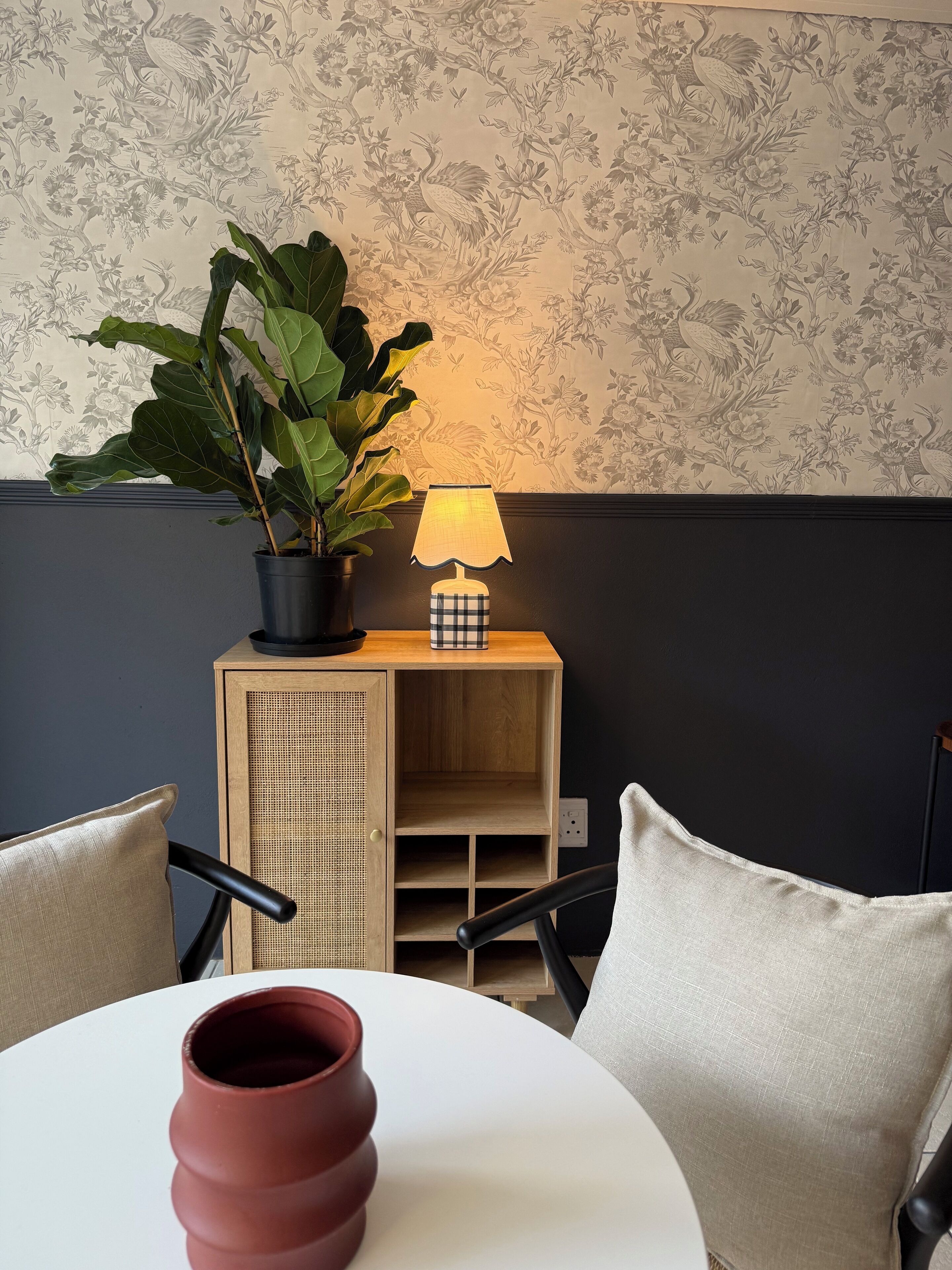 A room with a round table, two chairs, a potted plant, a lamp, and a cabinet with a woven front.