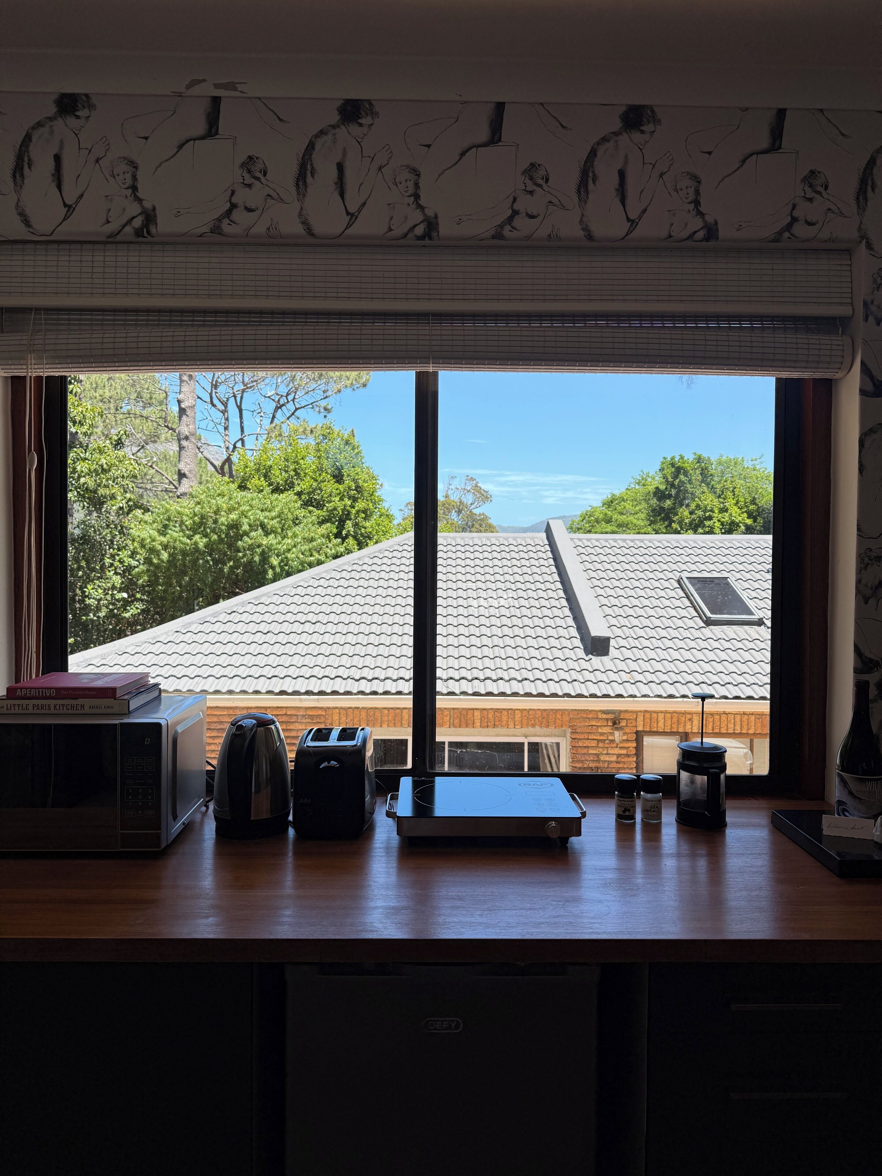 Ein Zimmer mit großem Fenster, das auf ein Haus mit Ziegeldach und Grünflächen blickt. Auf der Fensterbank stehen Küchengeräte, darunter eine Mikrowelle, ein Toaster und eine Kaffeemaschine.