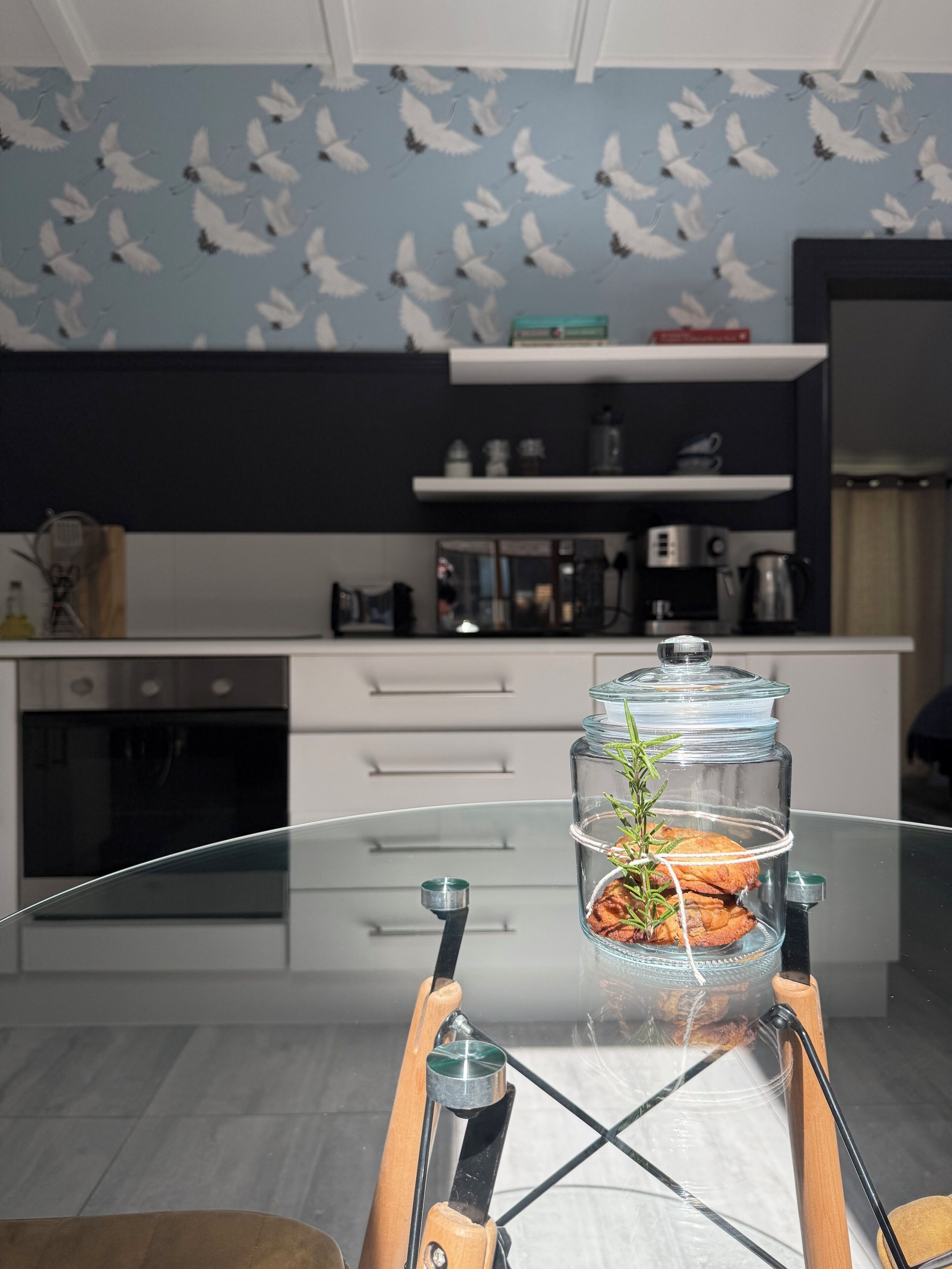 A modern kitchen with a glass-top table, a coffee machine, and a decorative jar with cookies and rosemary.