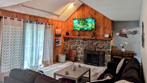 A cozy living room with a stone fireplace, wooden walls, a TV mounted above it, and a comfortable seating area with a sofa and a coffee table. - Modern Mountain Escape – Fun Filled Retreat! (Bartlett)