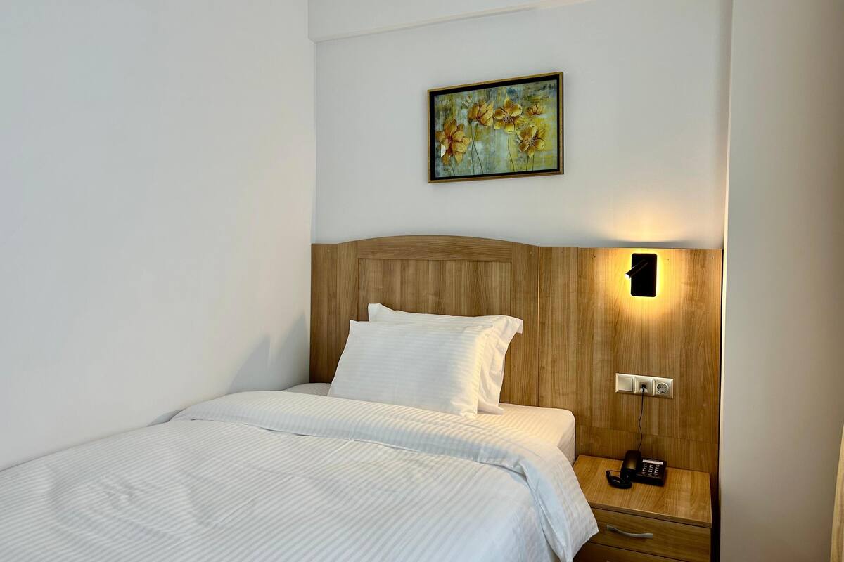 A neatly made bed with white linens, a wooden headboard, a bedside table with a lamp, and a painting on the wall.