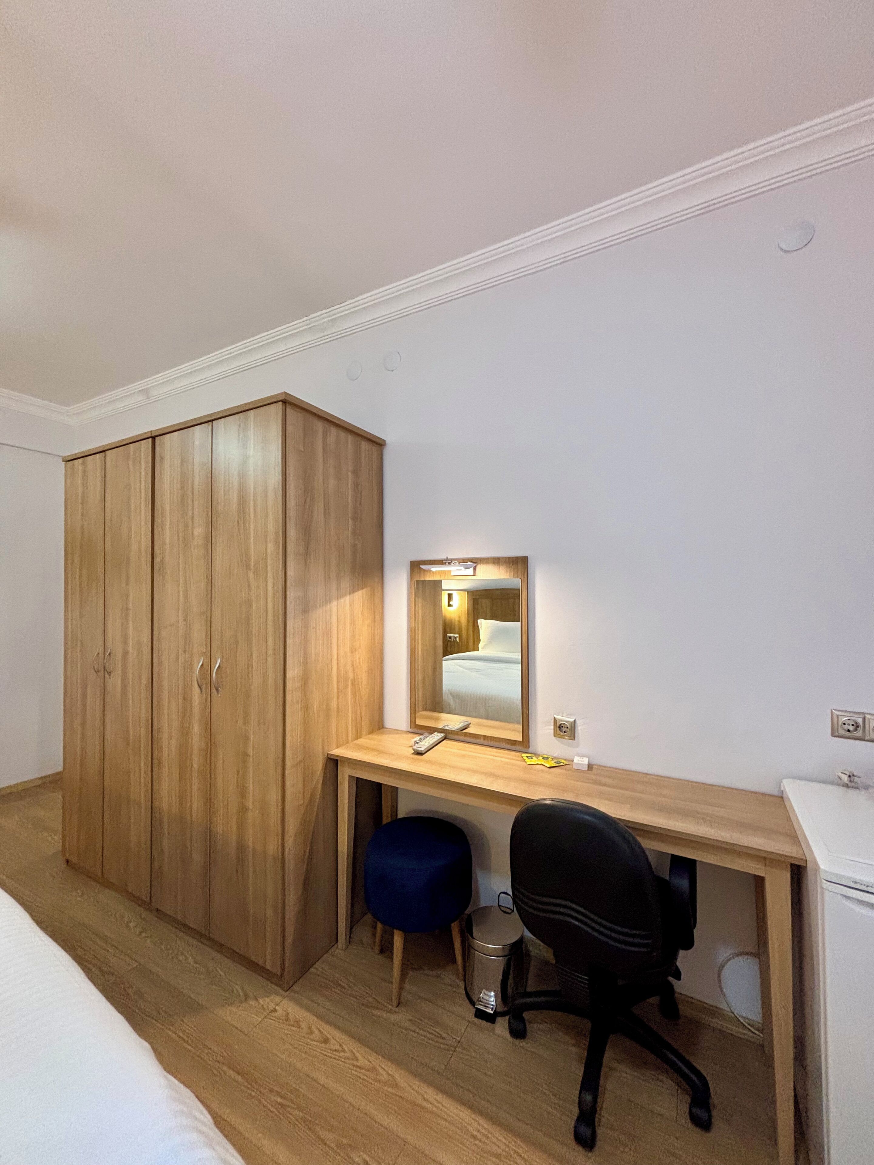 A bedroom with a wooden wardrobe, a desk with a mirror, a black office chair, and a small bedside table.