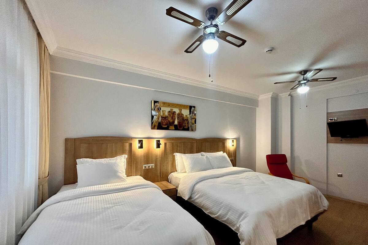 A hotel room with two beds, a ceiling fan, a mounted TV, and a painting on the wall.