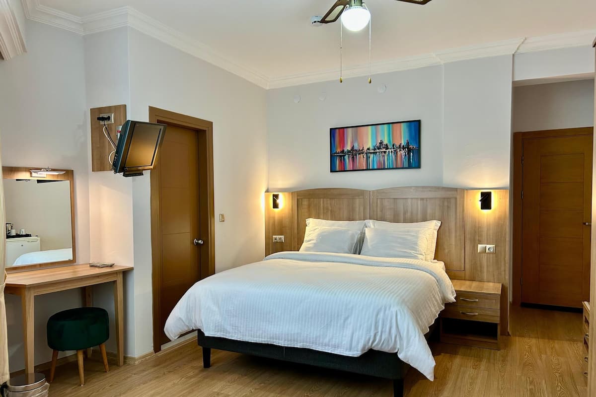 A hotel room with a bed, bedside tables, a desk, a chair, a mirror, a ceiling fan, and a painting on the wall.