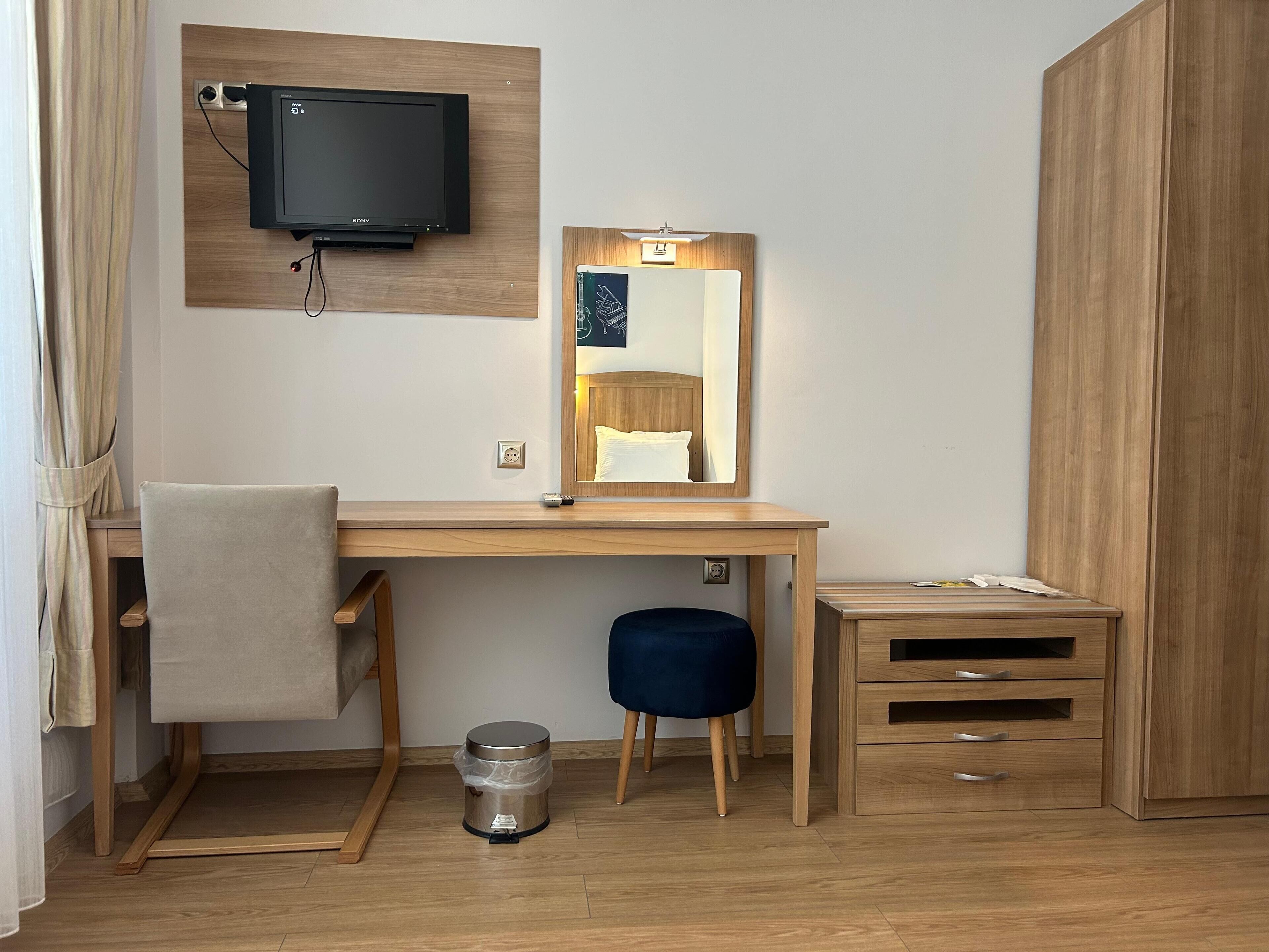 Una habitación de hotel con un escritorio de madera, un televisor montado en la pared, un espejo, un taburete pequeño, un cubo de basura y un armario.