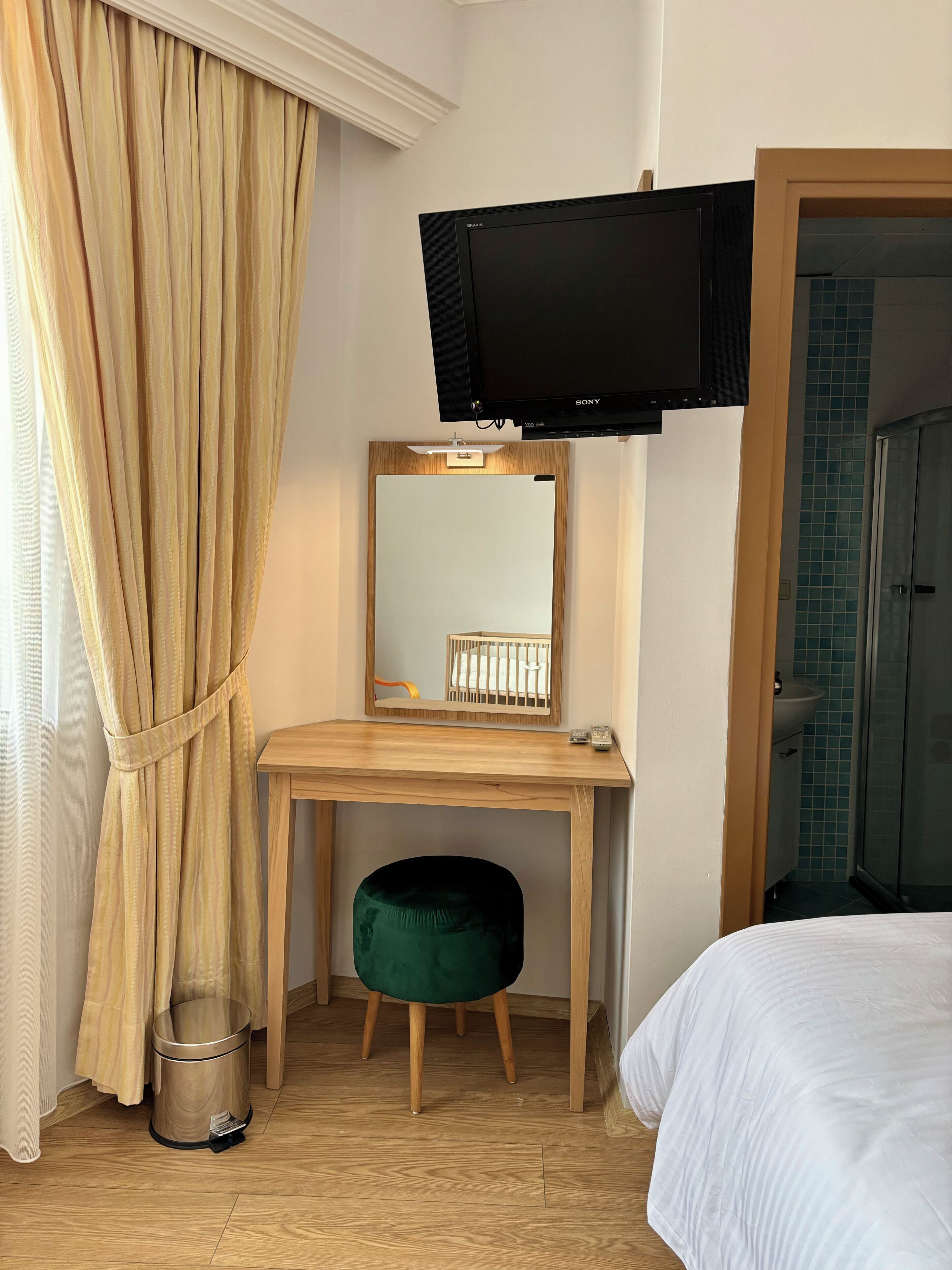 Habitación de hotel con escritorio de madera, televisor montado encima, taburete verde, cama, espejo, cubo de basura y zona de ducha.