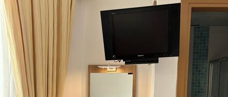 A hotel room with a wooden desk, a television mounted above it, a green stool, a bed, a mirror, a trash bin, and a shower area.