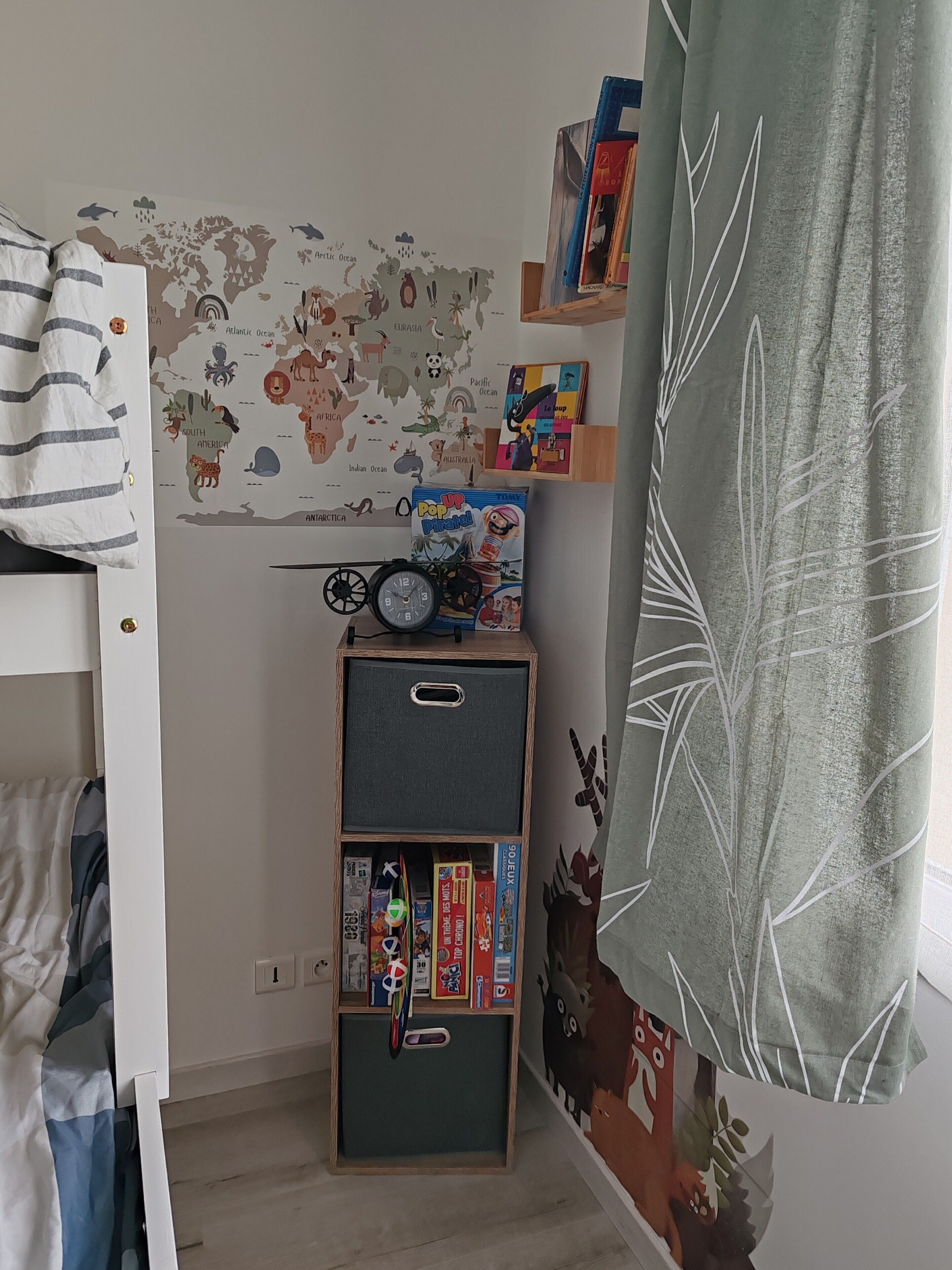 Quarto de criança com um mapa-múndi na parede, estante com livros e brinquedos, e cama com roupa de cama listrada.