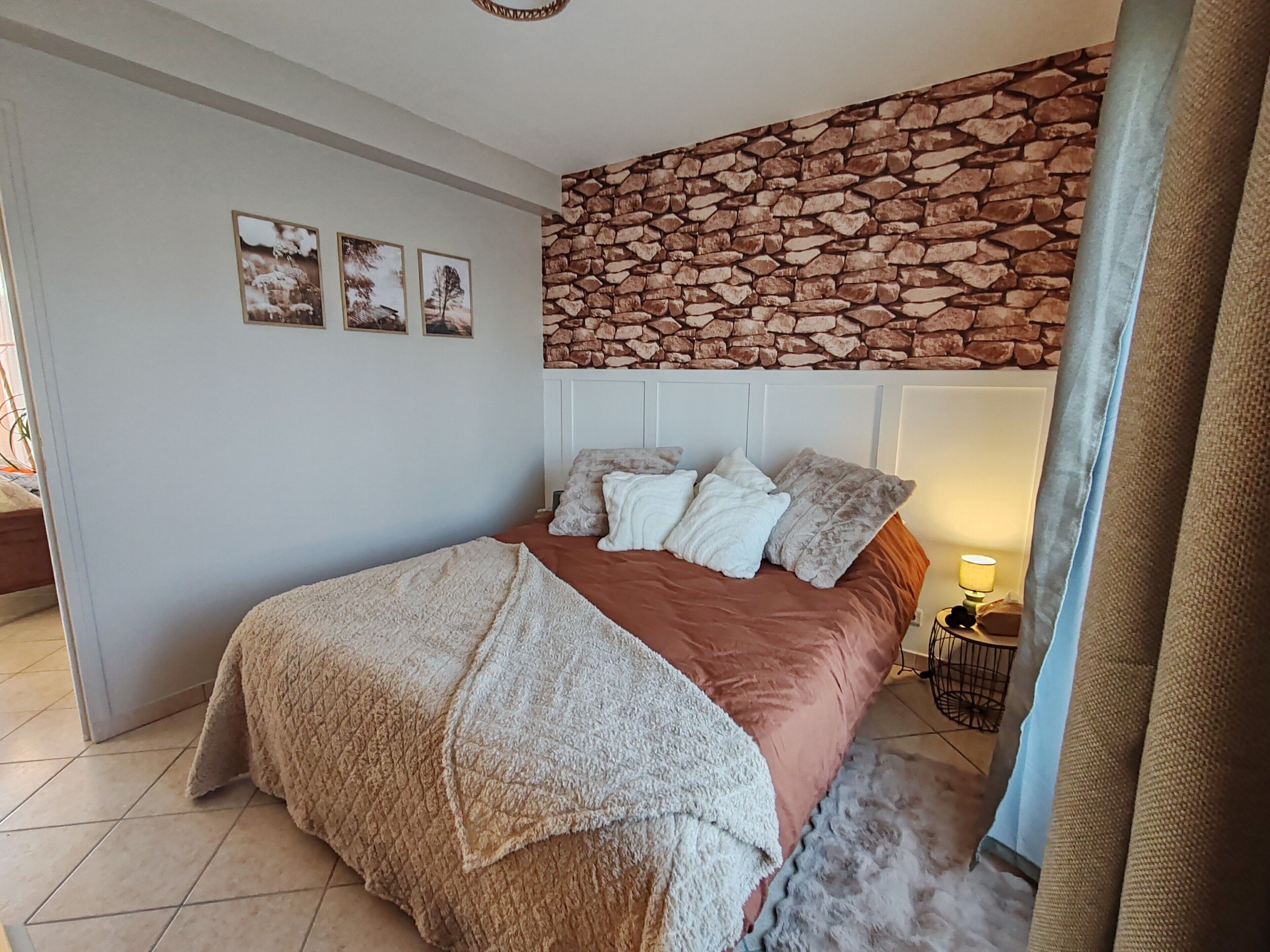 Un dormitorio con pared de piedra, una cama con muchos cojines, una lámpara de noche y una mesita con lámpara.