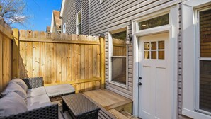 Une terrasse en bois avec un canapé et une petite table, une porte blanche et une fenêtre donnant sur la maison.