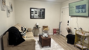 Una sala de estar con un sofá beige, una mesa de centro, un televisor montado en la pared y suelo de madera.