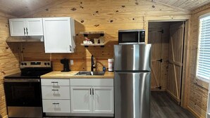 A compact kitchen with wooden walls, a stainless steel refrigerator, a microwave, a stove, and white cabinets. - Cozy cabin in Starkville (Starkville)