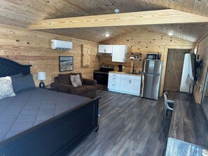 Una habitación compacta con cama, sofá, una pequeña cocina con refrigerador y televisión.