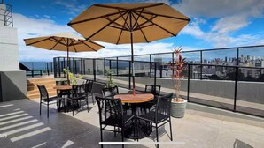 A rooftop terrace with outdoor seating, including tables and chairs, and large umbrellas. - Cozy and modern apartment with balcony. (João Pessoa)