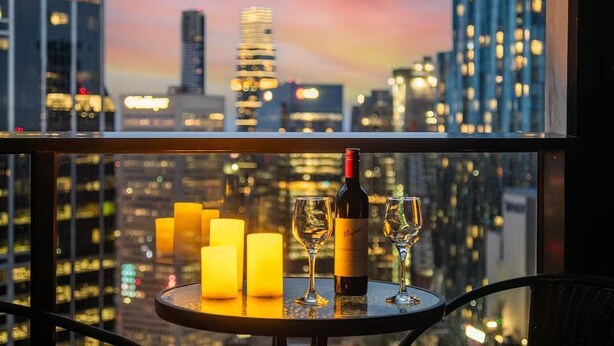 A table with a bottle of wine and two glasses, candles, and a cityscape view through a window. - Cozy 1BR Melbourne Escape | Balcony | Pool | GYM (Melbourne)