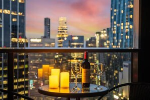 A table with a bottle of wine and two glasses, candles, and a cityscape view through a window. - Cozy 1BR Melbourne Escape | Balcony | Pool | GYM (Melbourne)