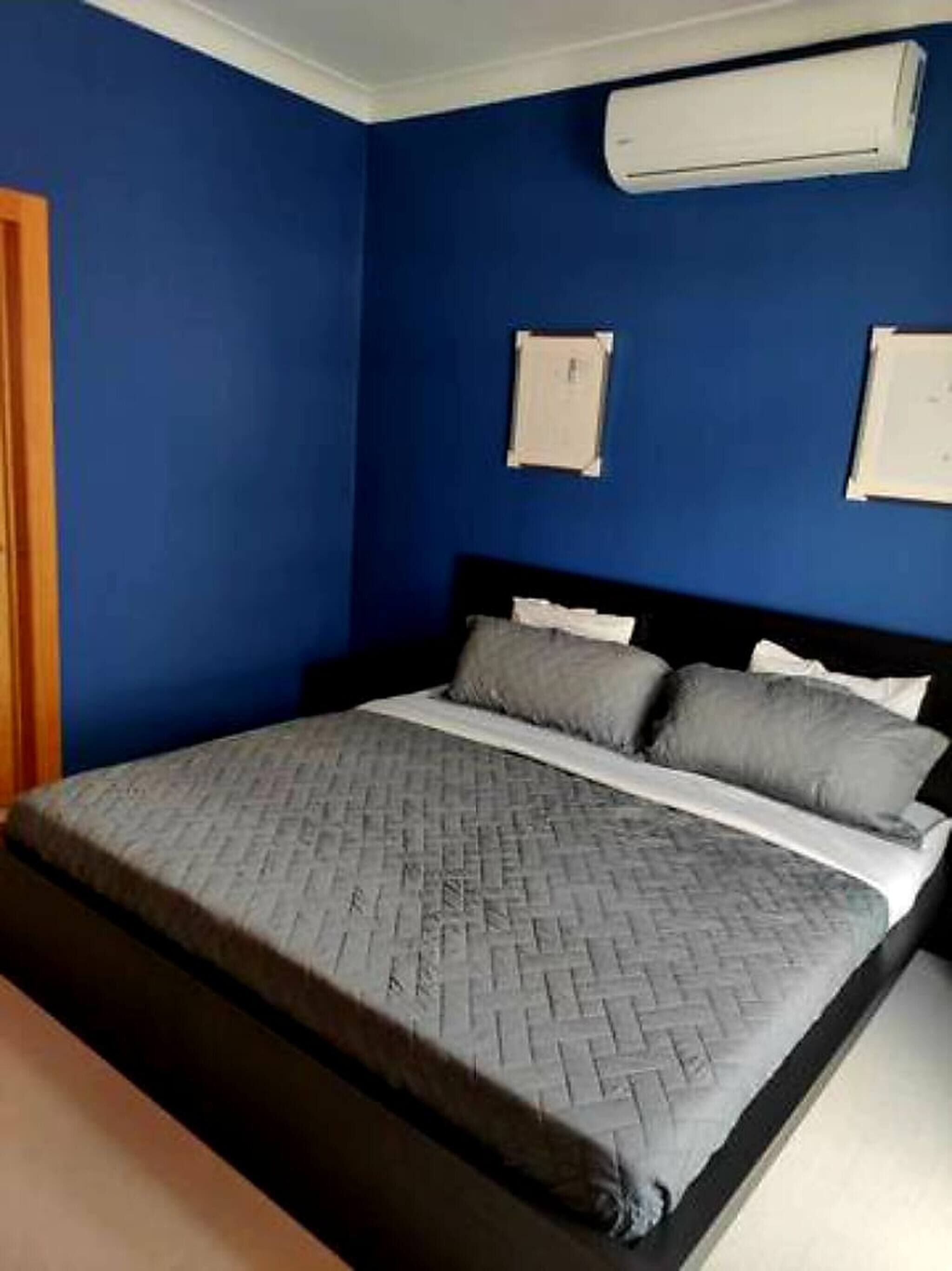 A bedroom with a blue wall, a bed with grey bedding, and two wall-mounted air conditioning units.