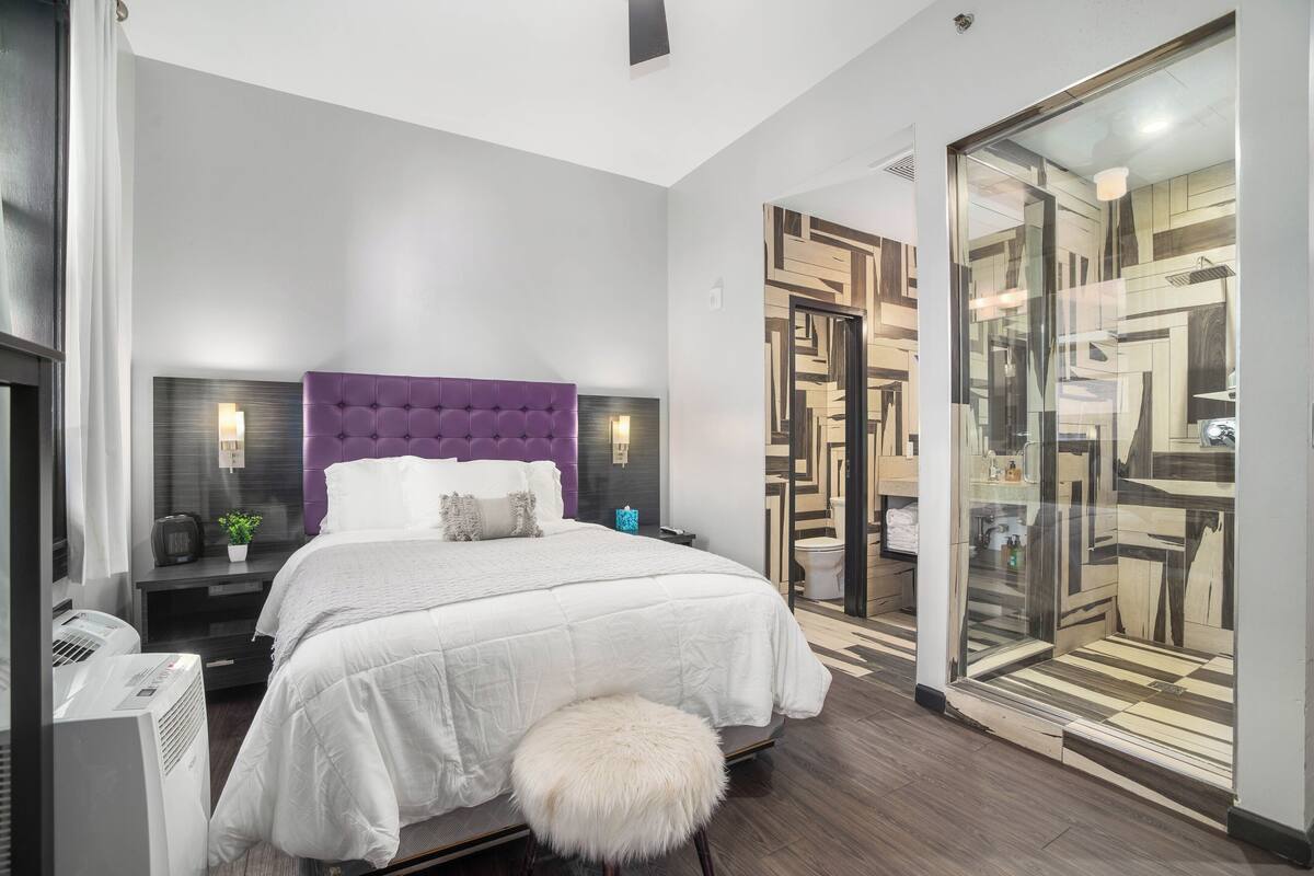A modern bedroom with a large bed, a purple headboard, bedside tables with lamps, and a glass shower enclosure.