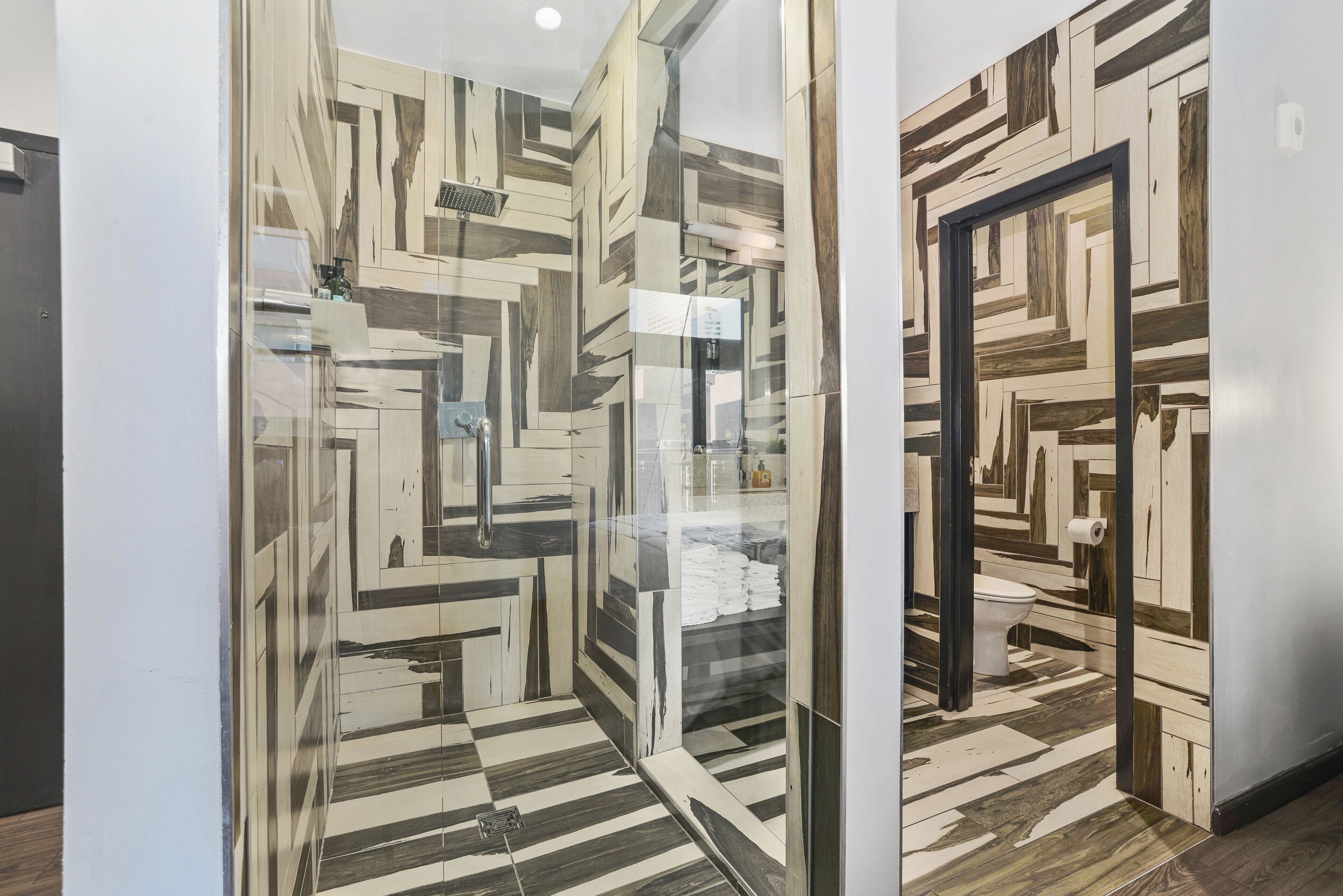 A modern bathroom with a geometric patterned floor and walls, a glass shower enclosure, and a visible toilet.