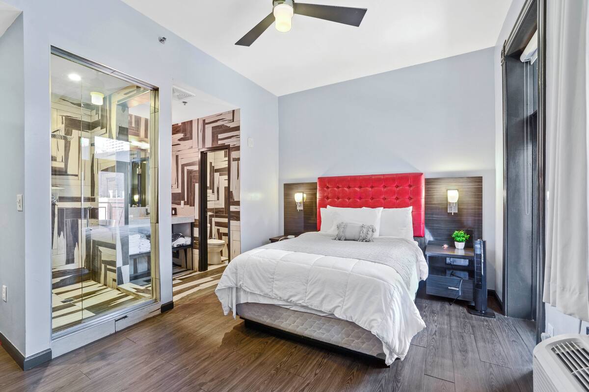 A modern bedroom with a large bed, a glass partition, a red headboard, and a bedside table with a lamp.