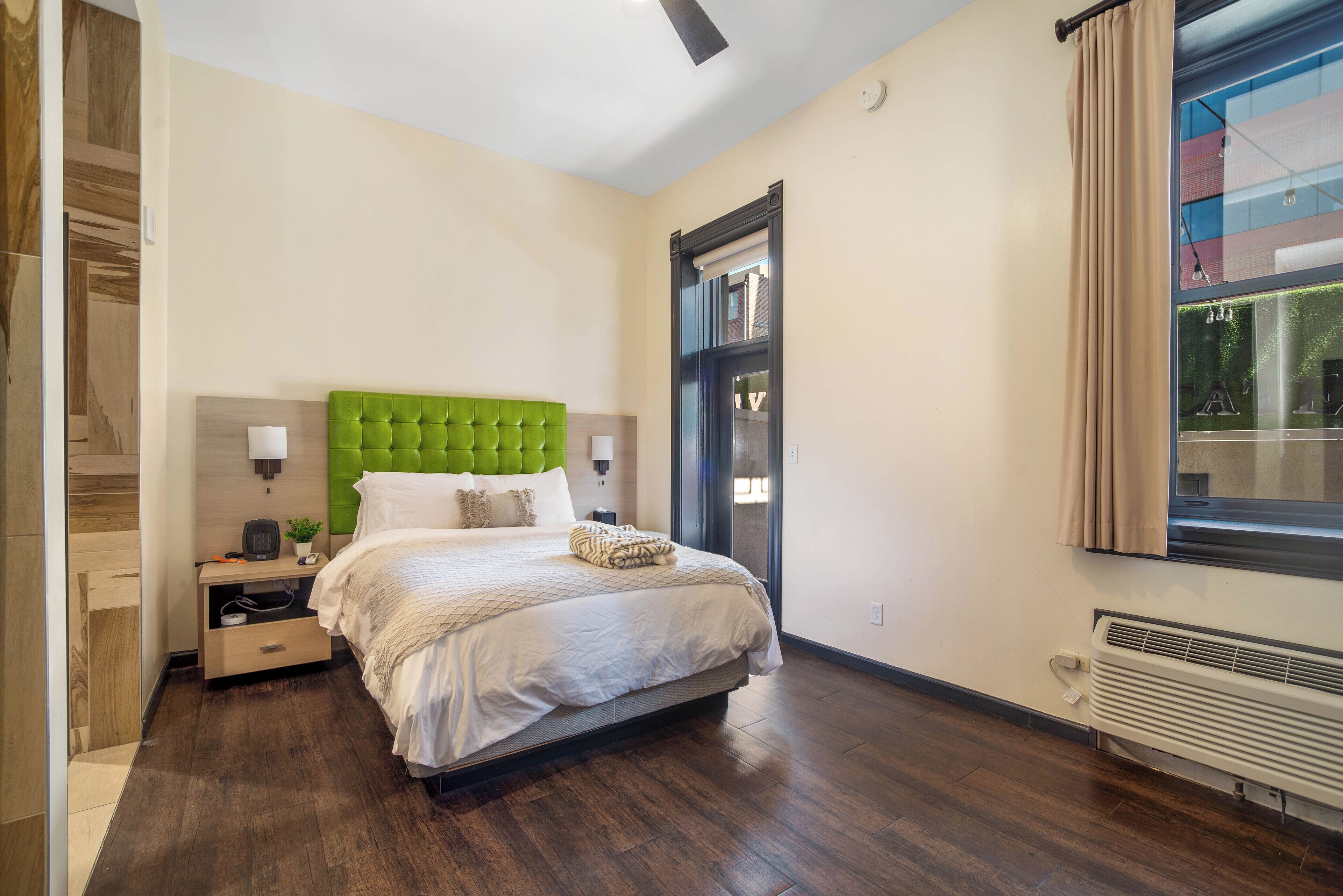 A bedroom with a green tufted headboard, a bed with white linens, a nightstand with a lamp, and a window with curtains.
