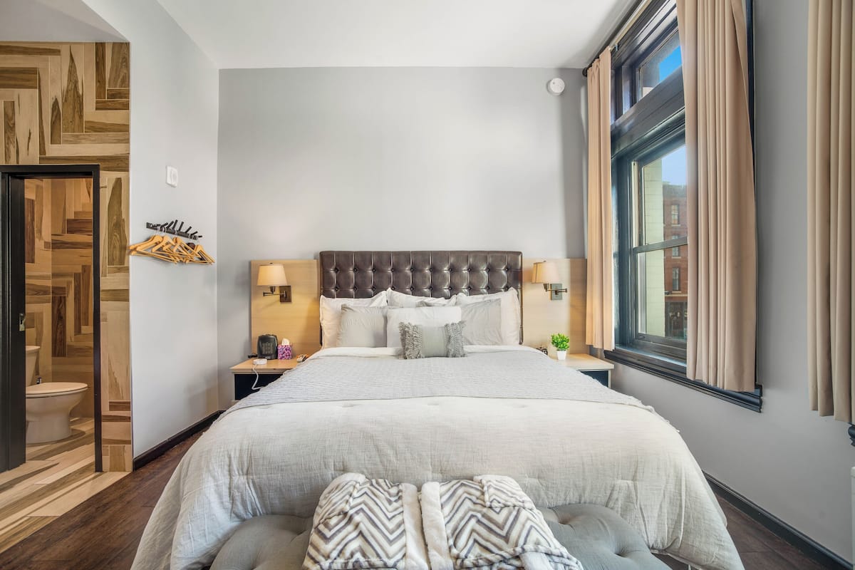 A bedroom with a tufted headboard, two bedside tables with lamps, a large bed with a grey and white striped blanket, and a window with beige curtains.
