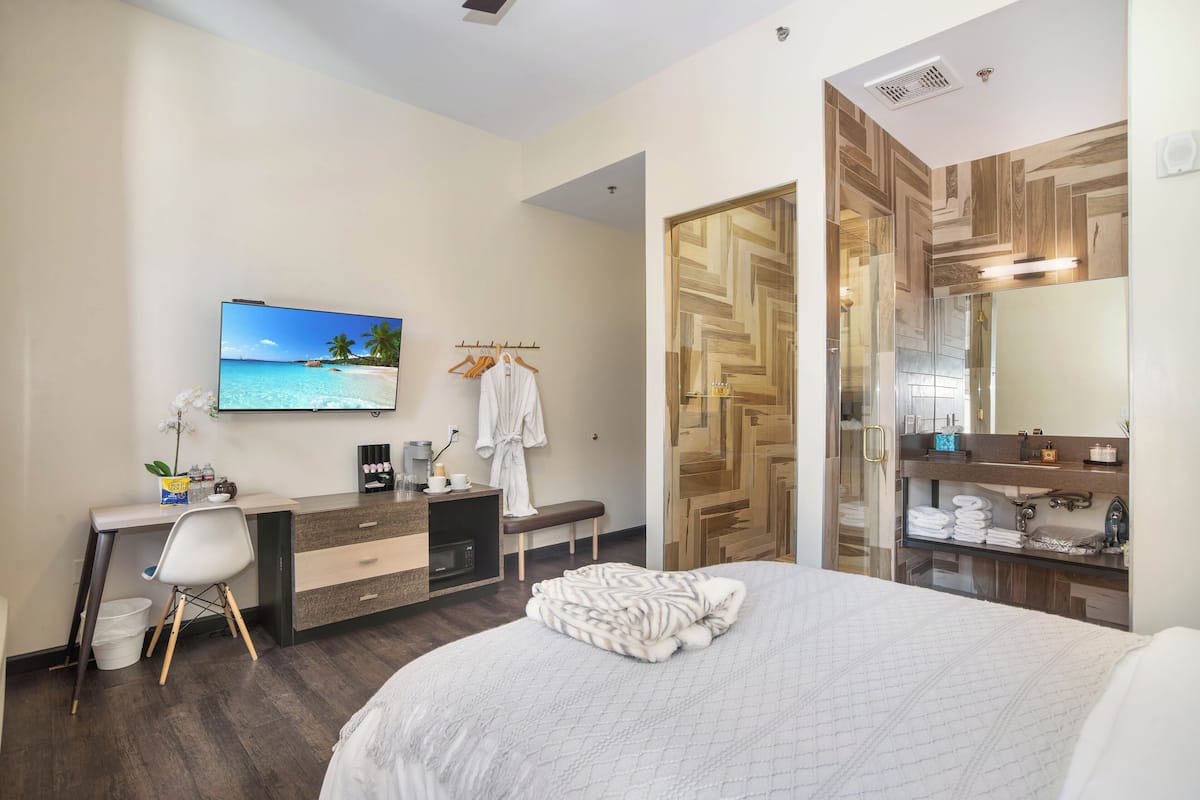 A hotel room with a bed, a desk with a computer, a TV, a robe, and a bathroom with a mirror and towel rack.