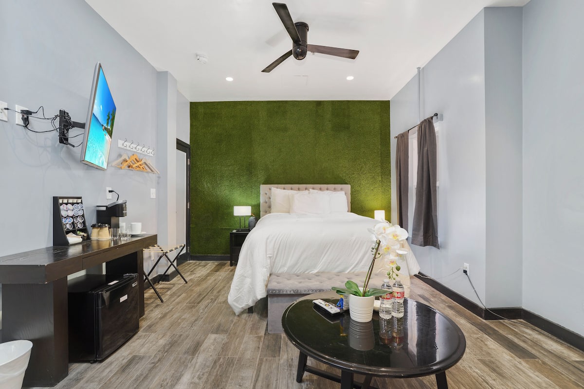A hotel room with a bed, a desk with a flat-screen TV, a ceiling fan, and a green accent wall.