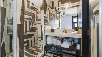 Et moderne badeværelse med et geometrisk flisemønster, et stort spejl og en vask med håndklæder og toiletartikler.