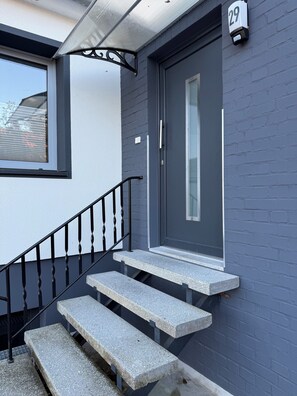Fachada moderna con una puerta gris, un cartel con el número 29 y unos pocos escalones de piedra que llevan a una puerta de cristal.