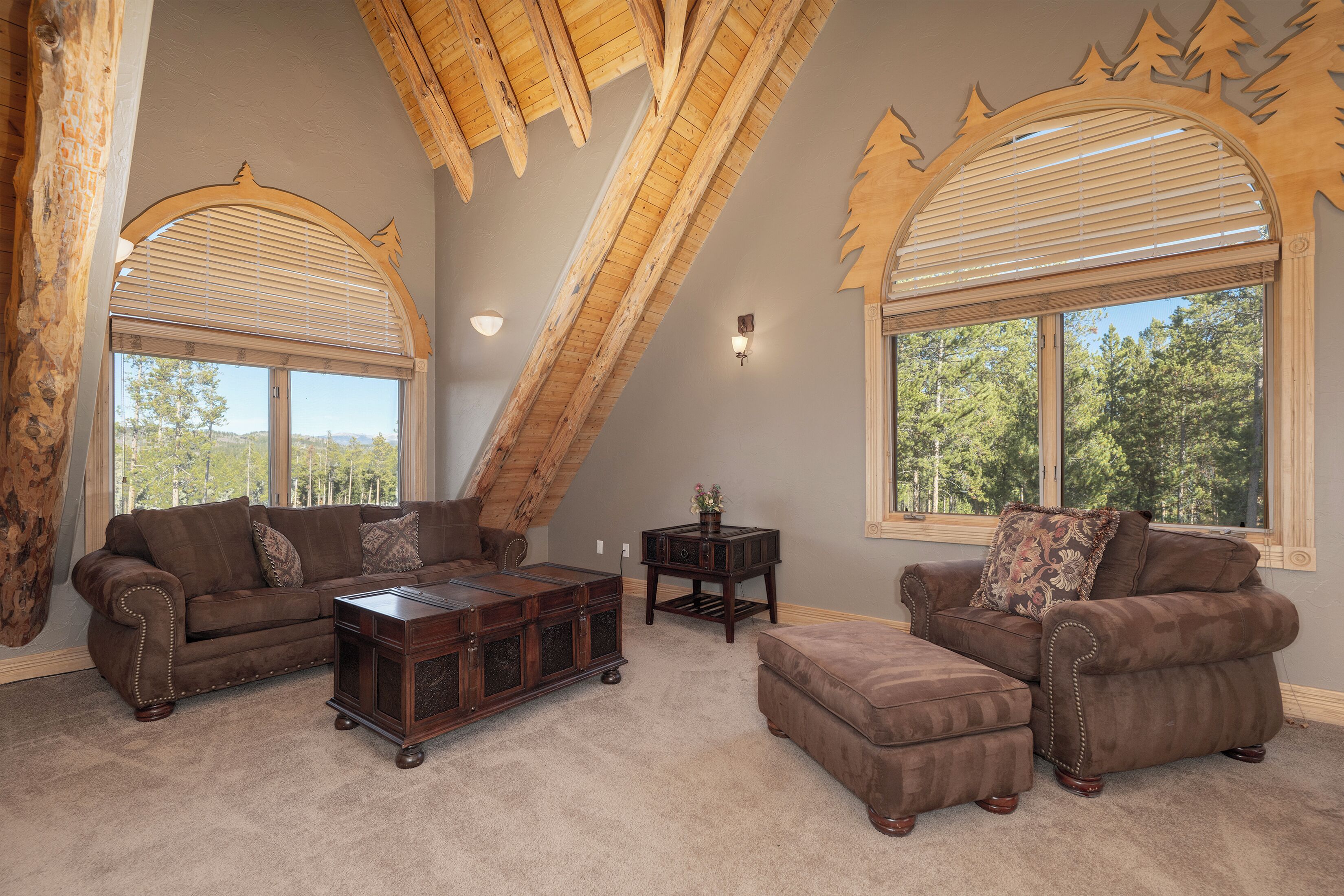 A spacious living room with high ceilings, wooden beams, and large windows offering a view of the outdoors.