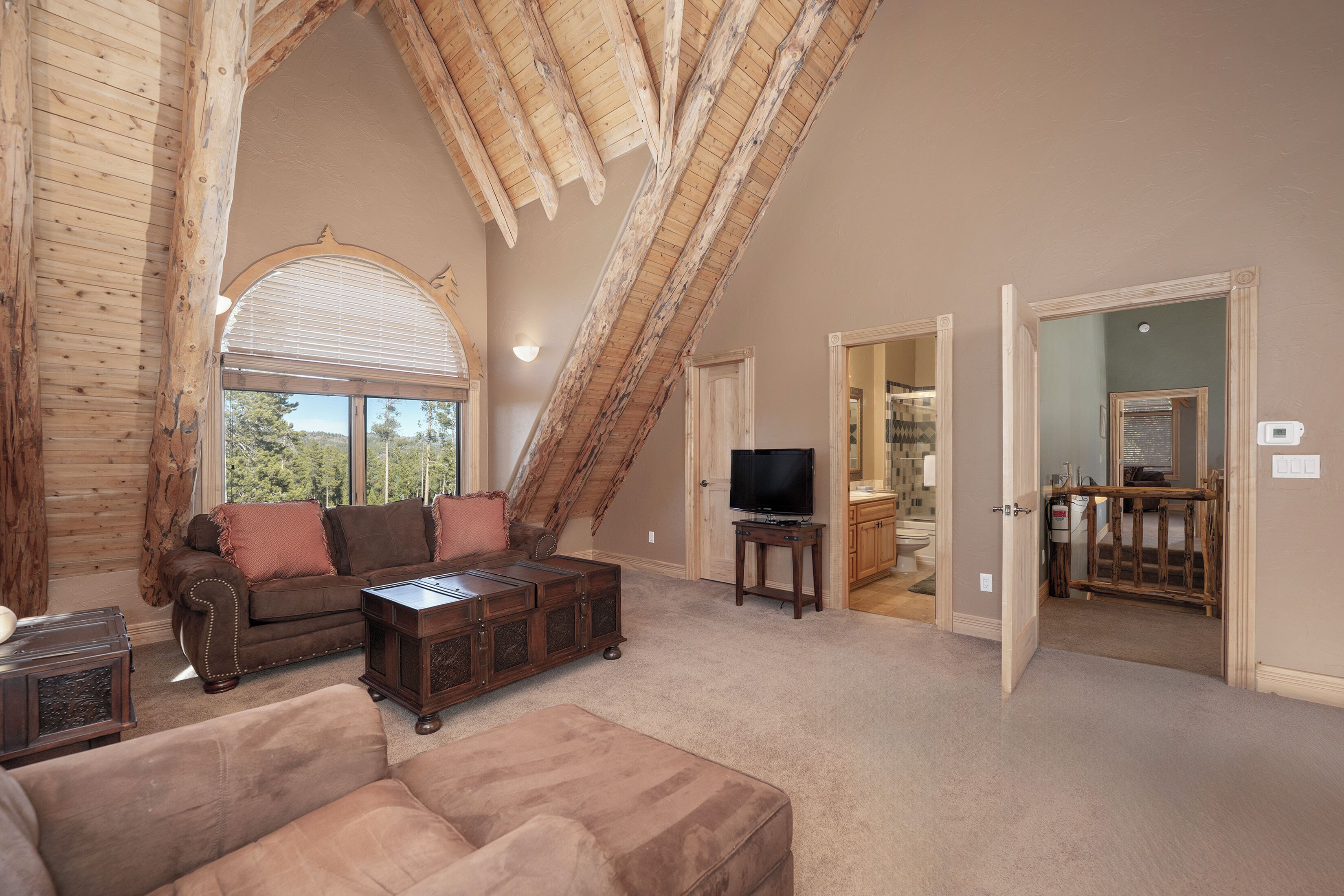 A spacious living room with a high ceiling, wooden beams, a large window, and a television on a stand.