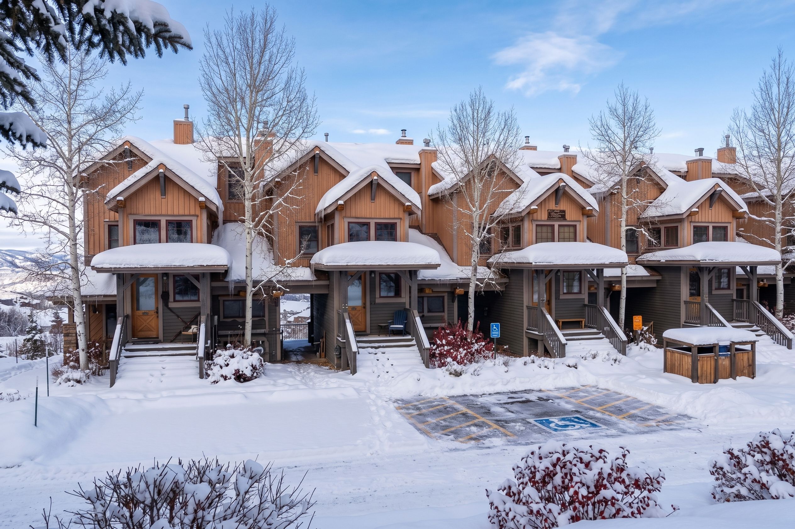 A row of wooden cabins with snow-covered roofs, a covered porch, and a parking area with a handicapped parking sign.