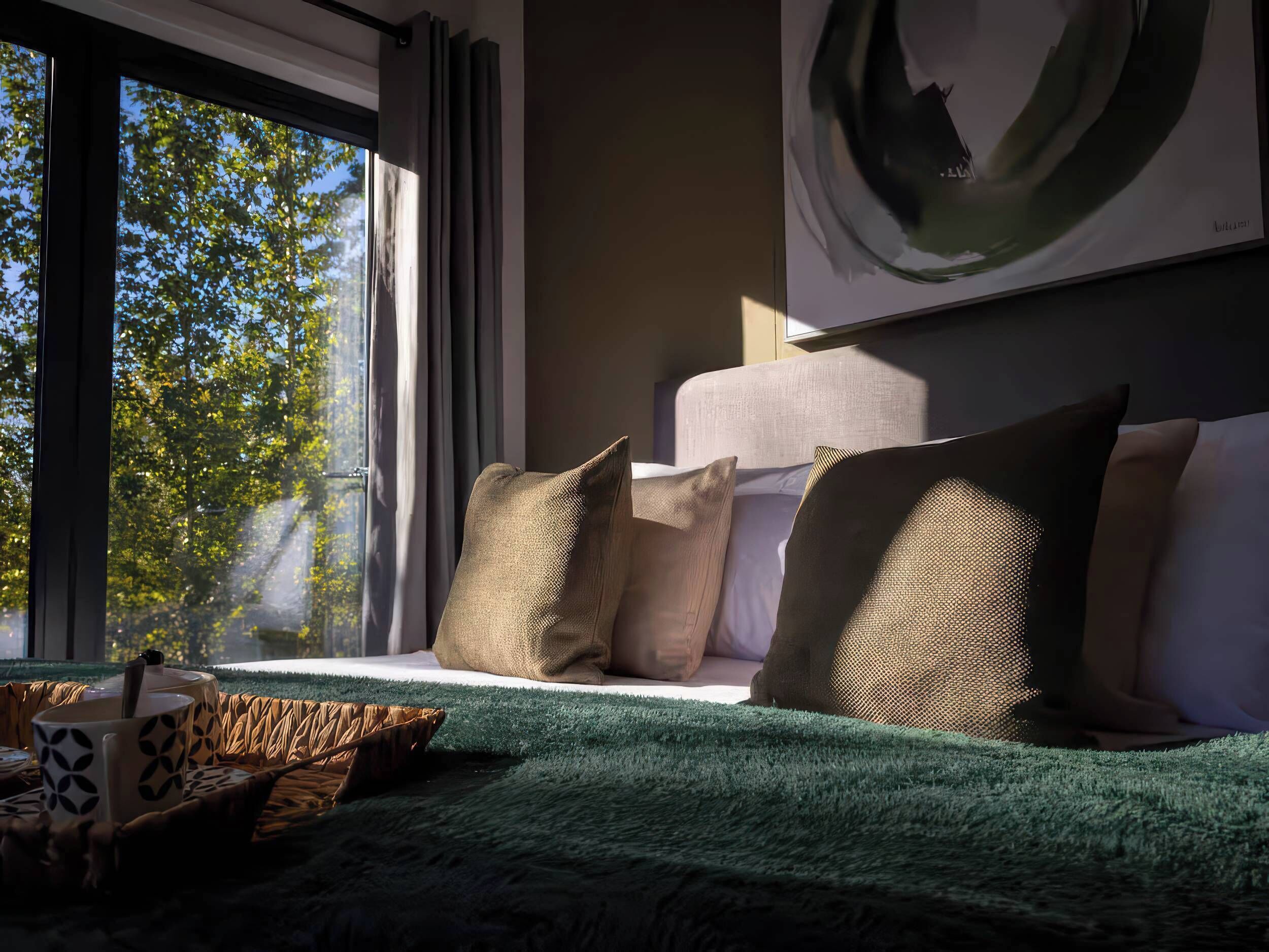A modern bedroom with a large window, a green textured bedspread, and a view of trees outside.