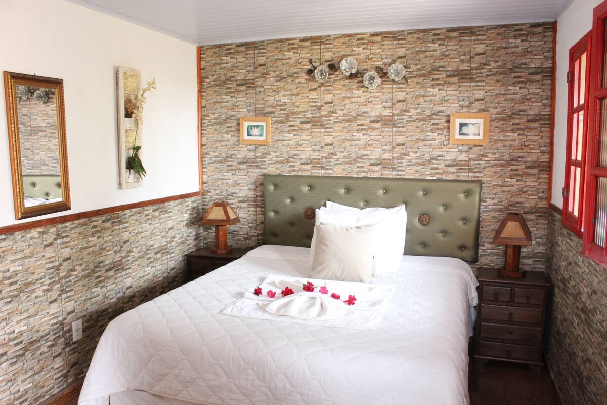 Een net opgemaakt bed met een gebordureerde witte deken, een bekleden hoofdbord in groen, en een decoratief kussen. Er zijn twee nachtkastjes met lampen, een muur met baksteenmotief, en ingelijste foto's aan de wand.