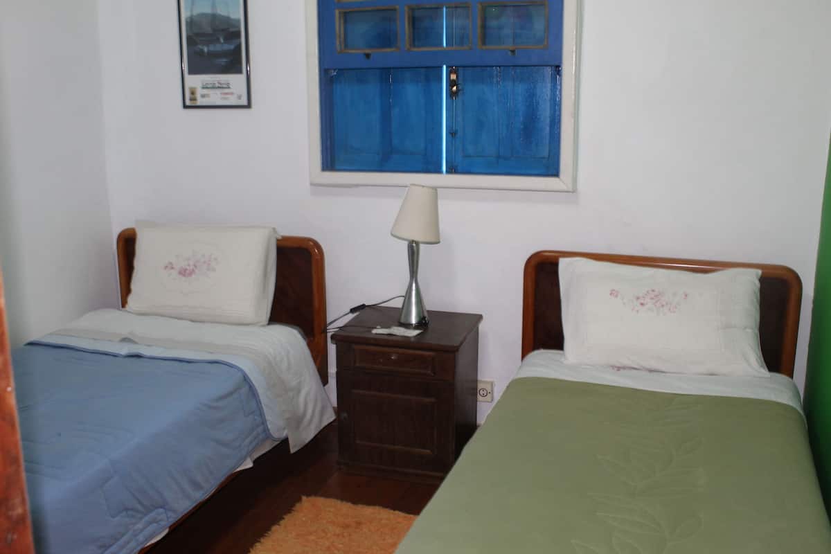 Een kamer met twee eenpersoonsbedden, een nachtkastje met een lamp, een raam met blauwe luiken en een ingelijst schilderij aan de muur.