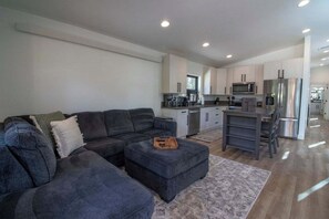 Ein modernes Wohnzimmer mit einem Ecksofa, einem kleinen Hocker und einer Küchenzeile mit Edelstahlgeräten.