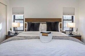 Ein ordentlich bezogenes Bett mit weißer Decke und zwei Kissen, ein Nachttisch mit Lampe sowie ein Fenster mit Jalousien.