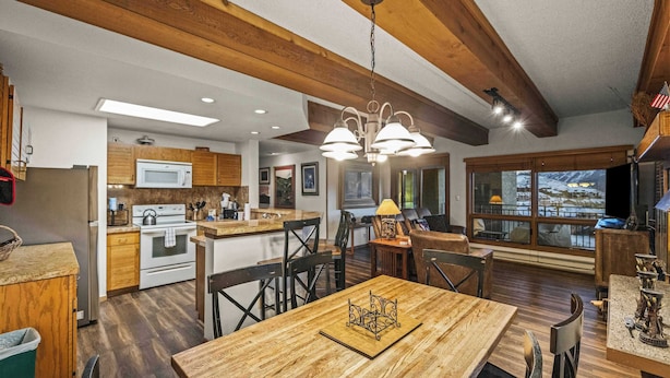 A spacious kitchen with wooden beams, a dining area with a chandelier, and a view of a snowy landscape through large windows. - Plaza 536 | Shuttle to Crested Butte Lifts | Hot Tubs+Sauna | 2BR Sleeps 6 (Crested Butte)