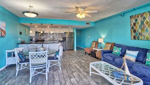 Ein modernes Wohnzimmer mit blauer Akzentwand, einem Essbereich mit weißen Stühlen, einem blauen Sofa mit dekorativen Kissen und einer Küche im Hintergrund.