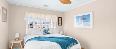 A bedroom with a bed, a ceiling fan, a window with curtains, a bedside table with a lamp, and a framed picture on the wall.