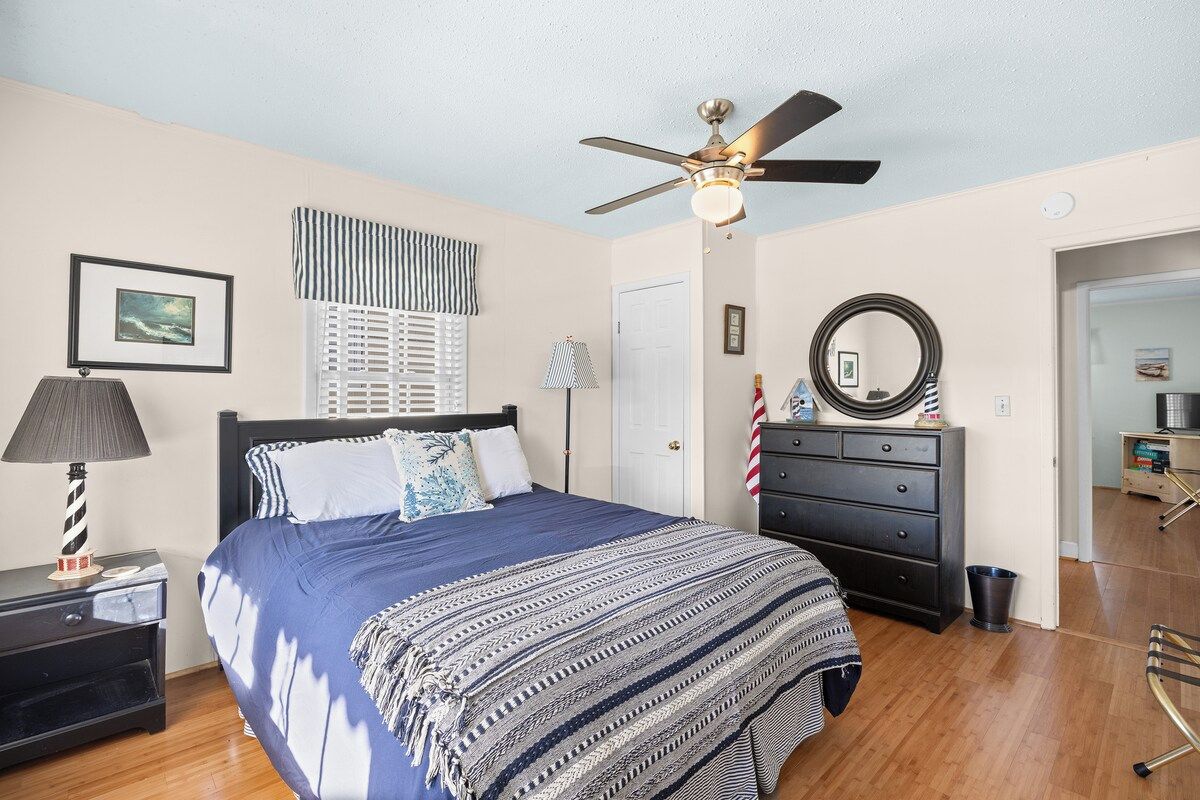 Un dormitorio con cama, ventilador de techo, cómoda, lámpara y un cuadro en la pared.