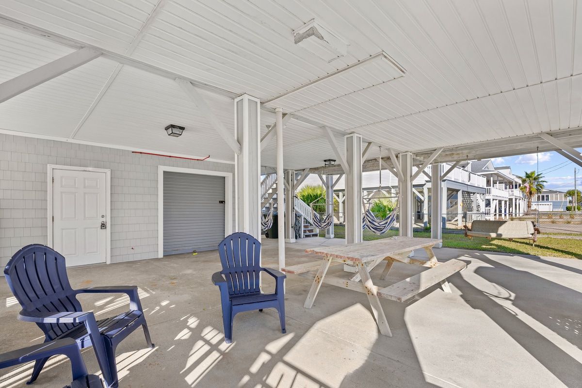 A covered outdoor area with two Adirondack chairs, a concrete floor, and a white building in the background.