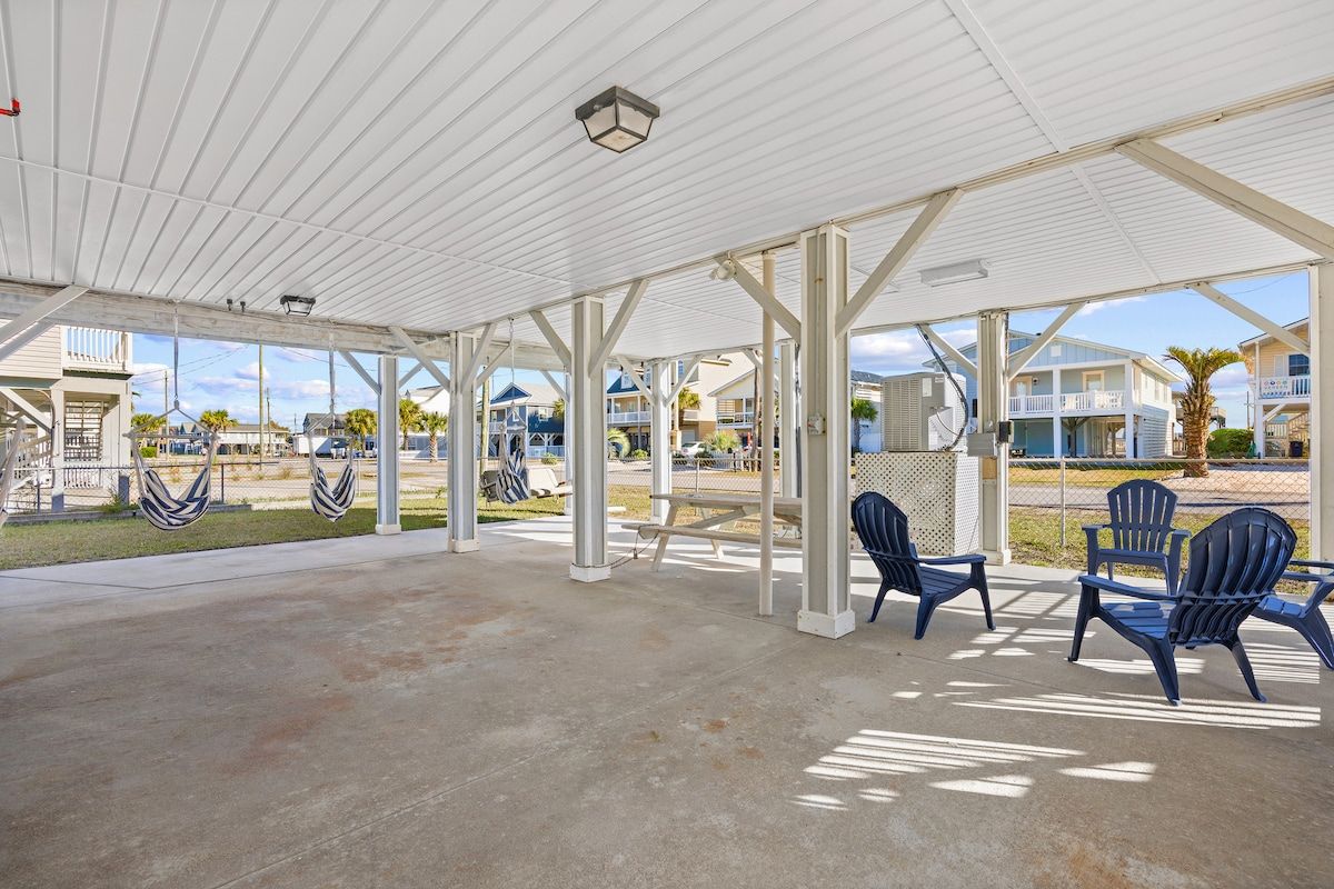 A covered outdoor area with a concrete floor, white metal beams, and blue Adirondack chairs.