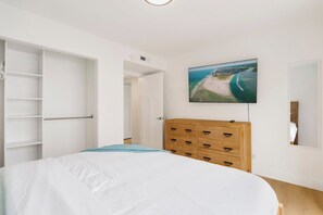 Un dormitorio con una cama, un aparador de madera, un armario y un televisor de pared mostrando una vista aérea de una playa.