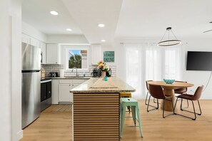 A modern kitchen with a central island, a dining area with a round table and chairs, and a flat-screen TV mounted on the wall.