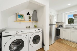 Zona de bugaderia amb dues rentadores Samsung, una petita estant amb objectes decoratius, i una cuina amb armaris blancs i un frigorífic d'acer inoxidable.