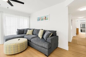 A modern living room with a grey sectional sofa, a patterned ottoman, and a ceiling fan.