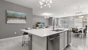 A modern kitchen with a large island, stainless steel appliances, and a dining area with a view of the outdoors. - Upstay Resort Villa - Pool, Hot Tub, Sleeps 27 (Kissimmee)