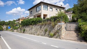 Una casa de dos pisos con un muro de piedra y un portón de metal, situada al lado de una carretera.